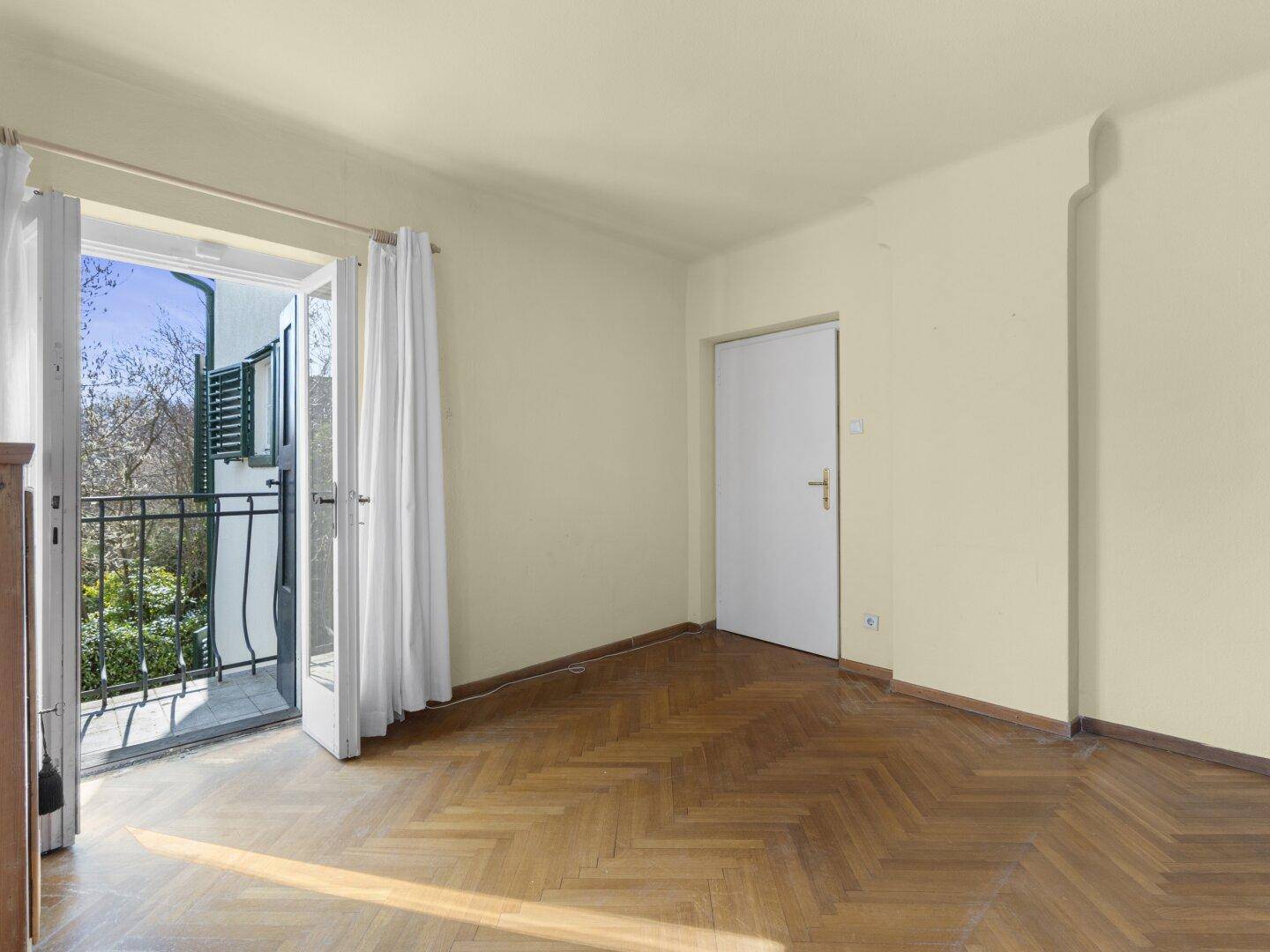 Leerer Raum mit Fischgrätparkett und Zugang zu einem kleinen Balkon.