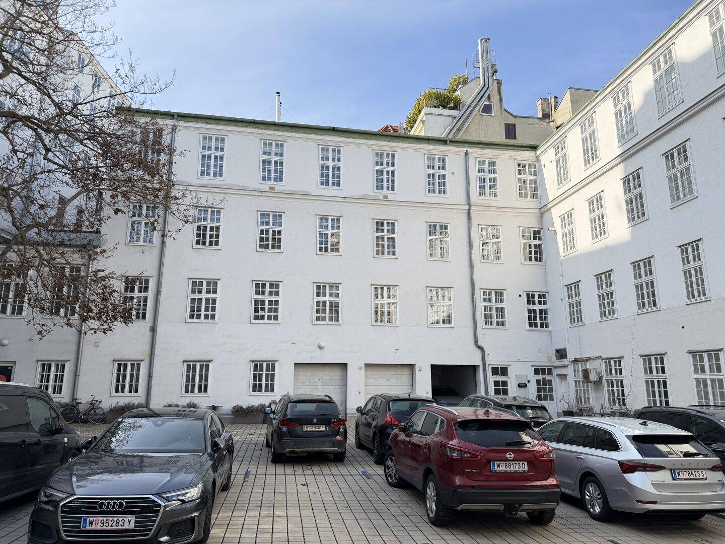 Weitläufiger Innenhof mit parkenden Autos vor einer historischen weißen Gebäudefassade mit vielen Fenstern.