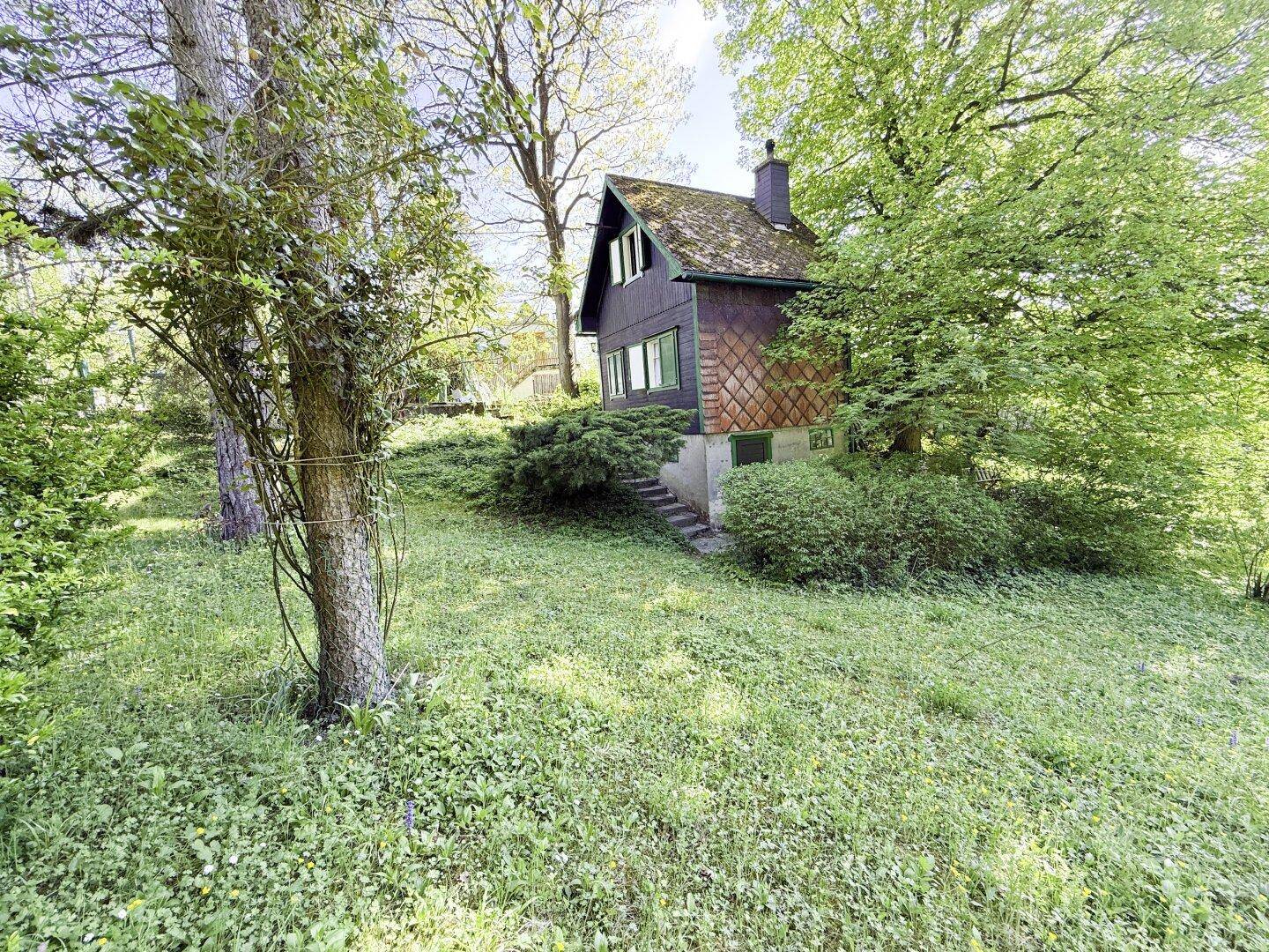 Kleines, rustikales Haus mit dunkler Holzfassade und grünen Fensterläden, umgeben von einem naturnahen Garten.