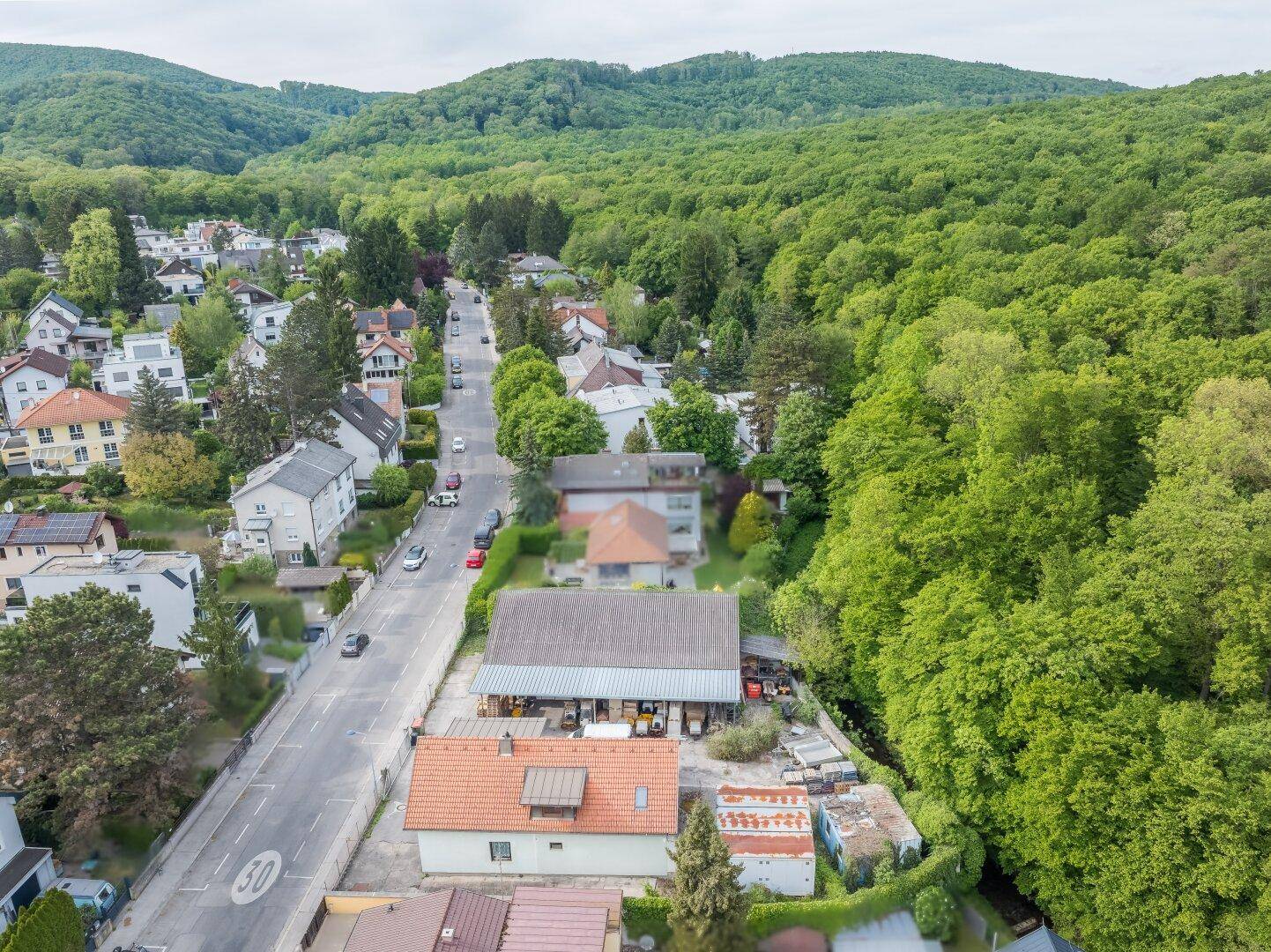 Luftbild einer Straße durch ein Wohngebiet, umgeben von dichtem Wald und grünen Hügeln.