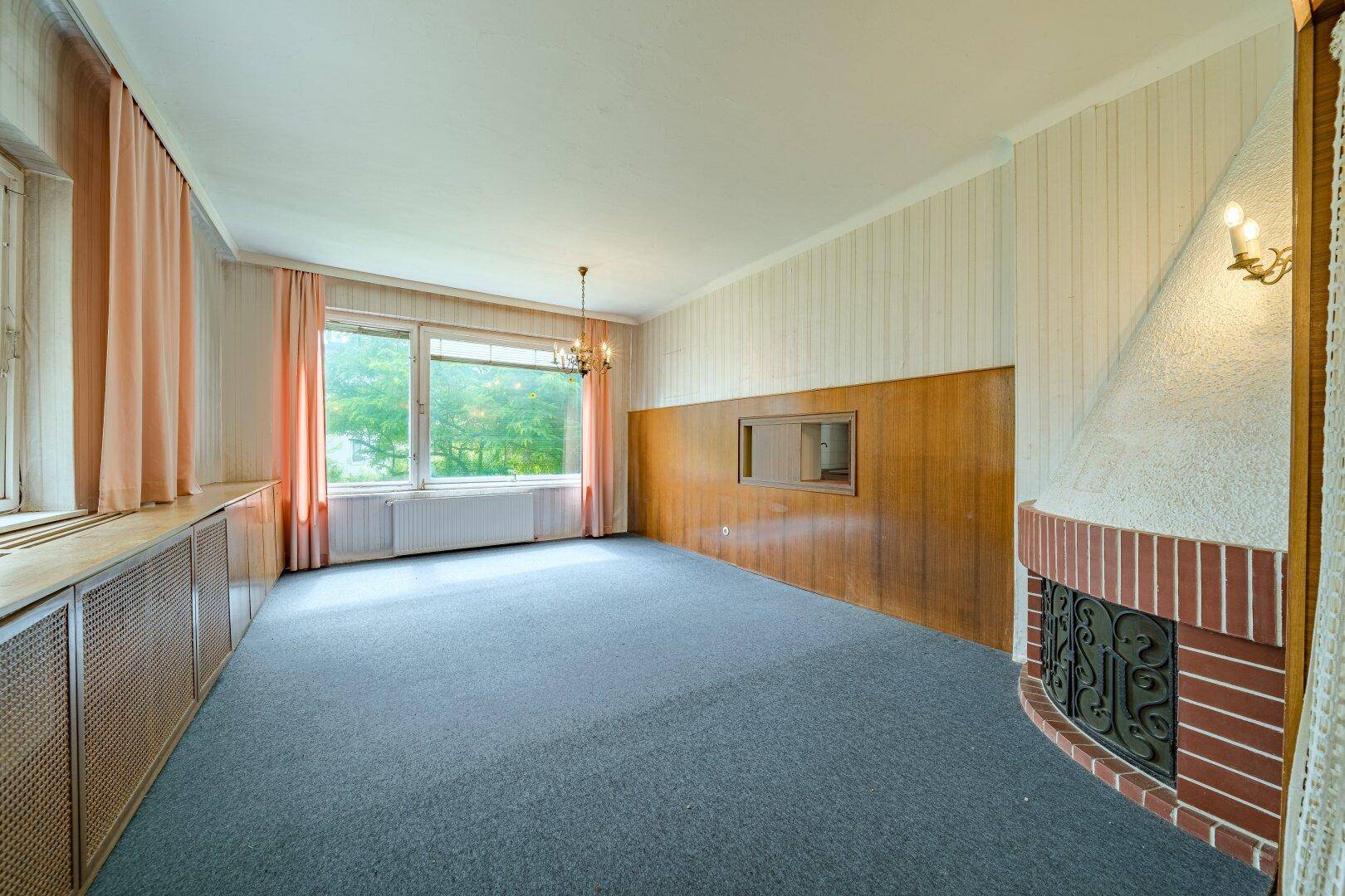 Helles Wohnzimmer mit Kamin, großen Fenstern und Teppichboden, bereit zur Renovierung.