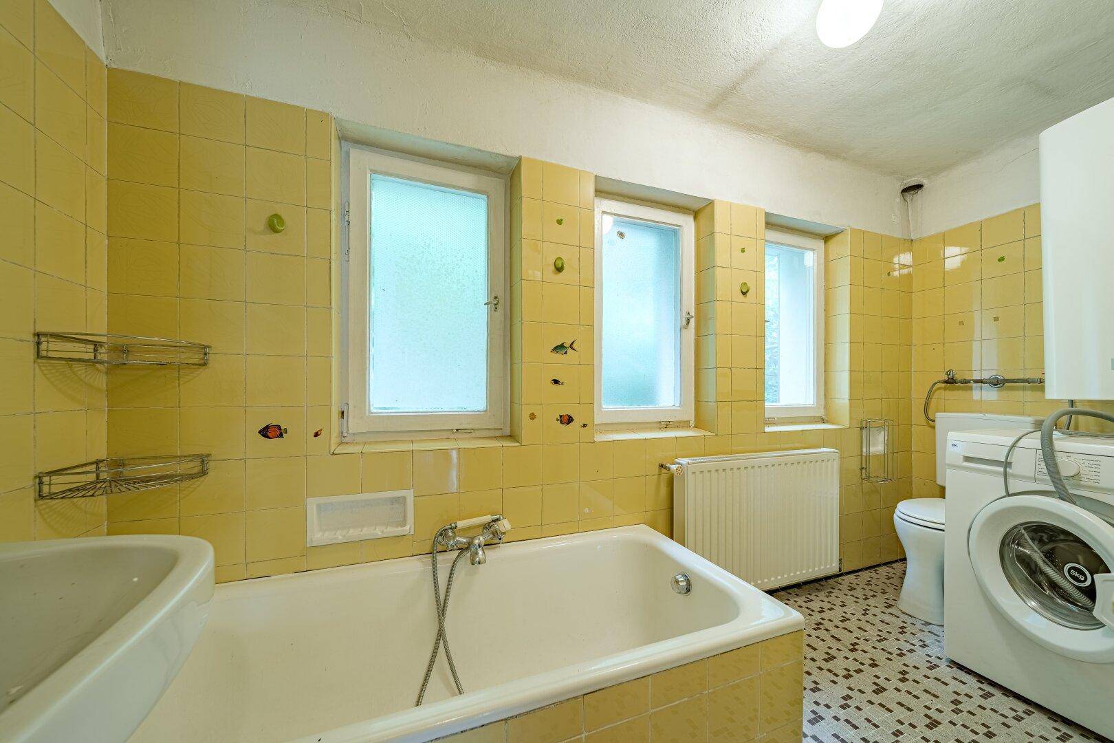 Funktionales Badezimmer im Stil der 80er Jahre mit gelben Fliesen, Badewanne, Toilette und Waschmaschine.
