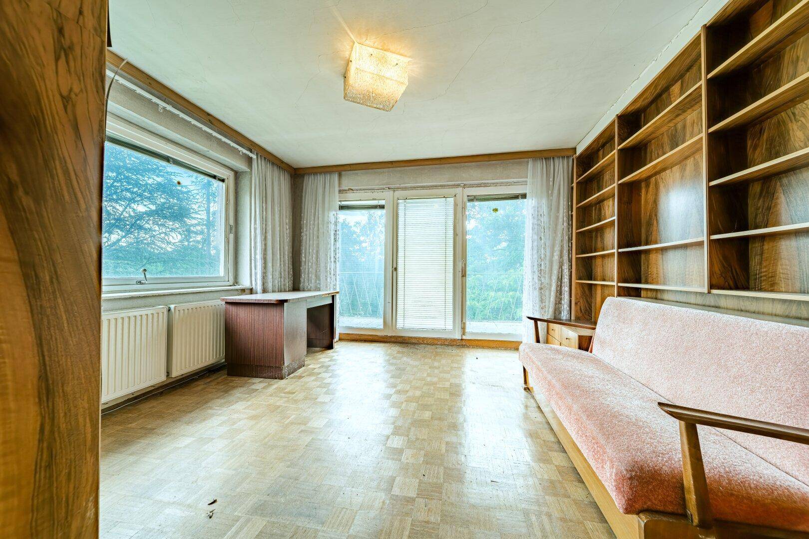Raum mit Parkettboden, großem Fenster, Zugang zum Balkon und eingebautem Bücherregal.