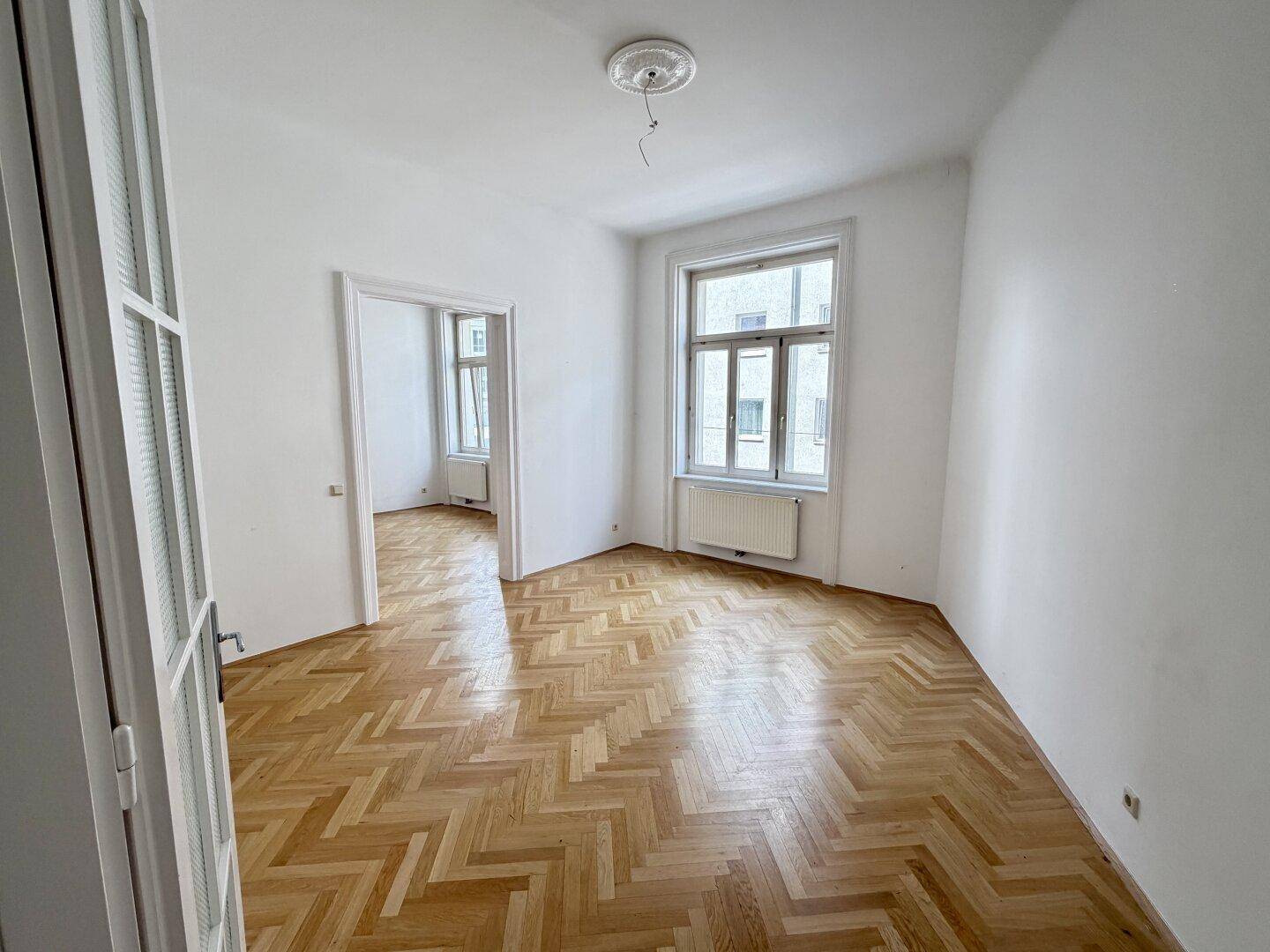 Geräumiger Wohnbereich mit klassischem Fischgrätparkett und Blick durch eine offene Tür in einen weiteren Raum.
