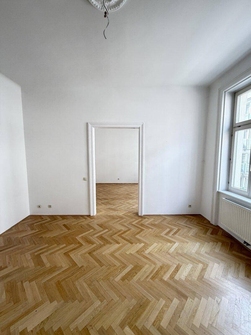 Leerer Raum mit Fischgrätparkett und einer breiten Türöffnung, die zu einem angrenzenden Zimmer führt.