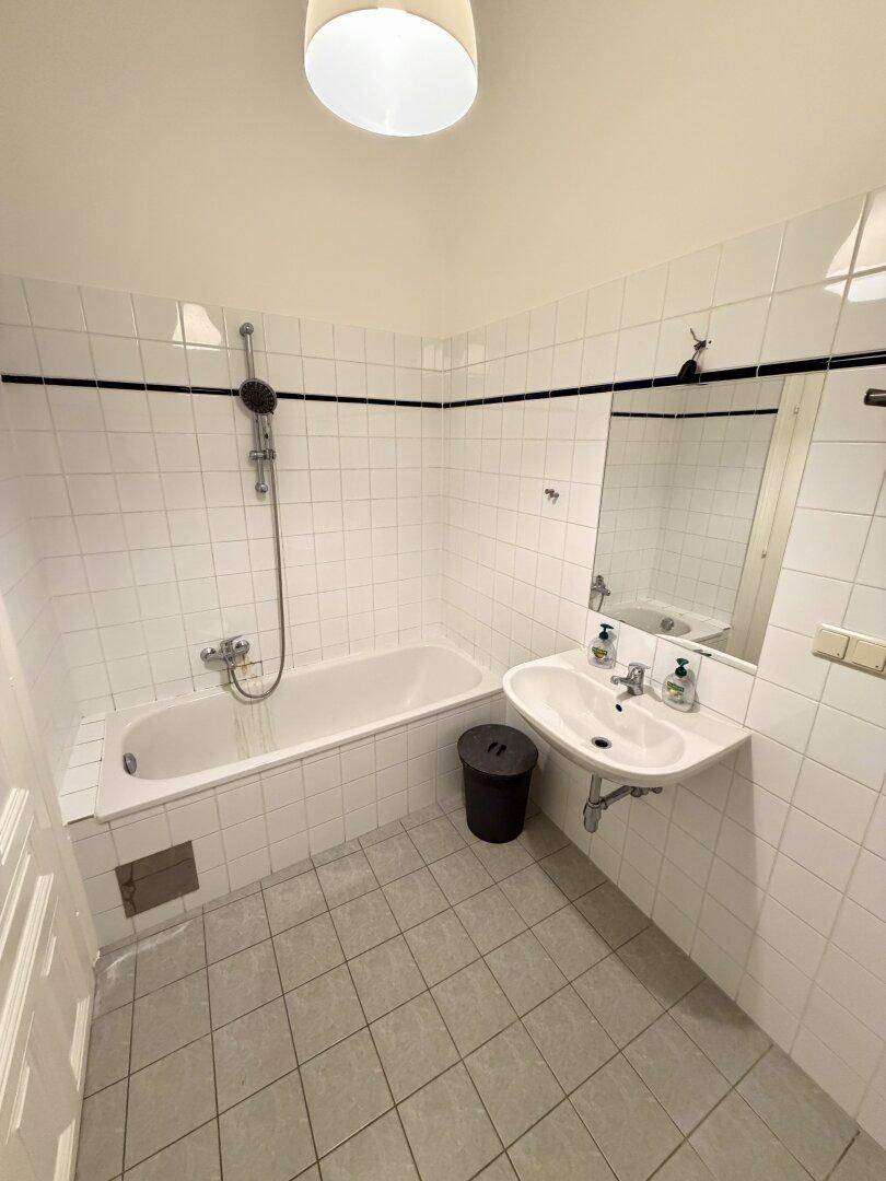 Helles Badezimmer mit Badewanne, Duscharmatur, Waschbecken und weißen Fliesen an Wänden und Boden.