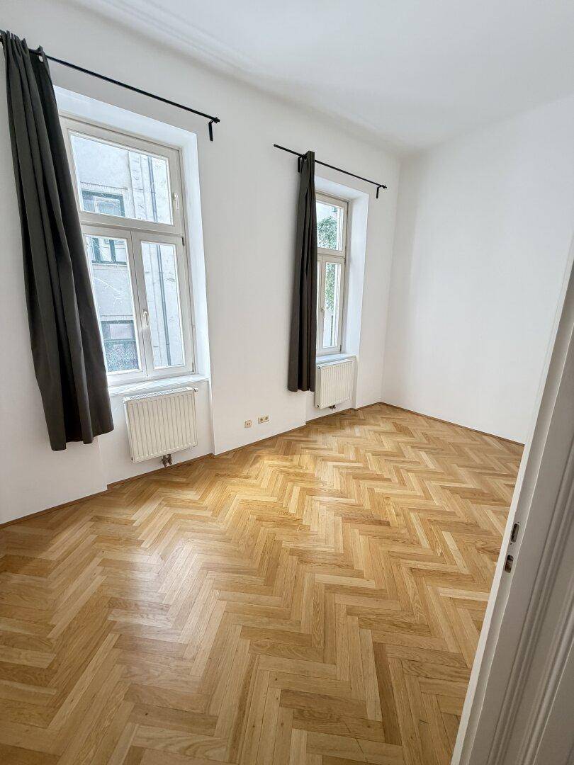 Schlafzimmer mit Fischgrätparkett, zwei Fenstern und dunklen Vorhängen, die für Privatsphäre sorgen.
