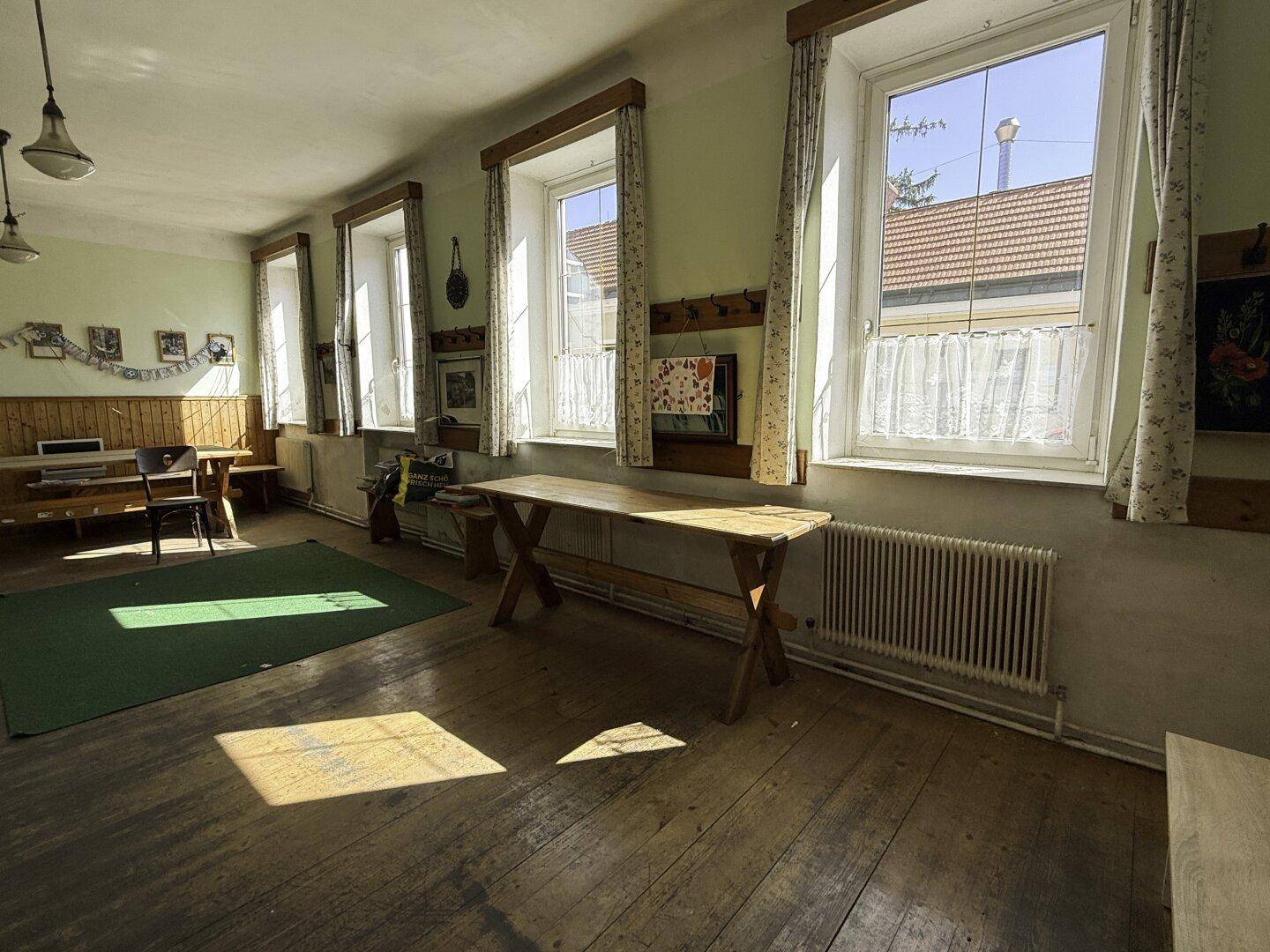 Geräumiger Gemeinschaftsraum mit Holzböden, großen Fenstern und einfacher Einrichtung.