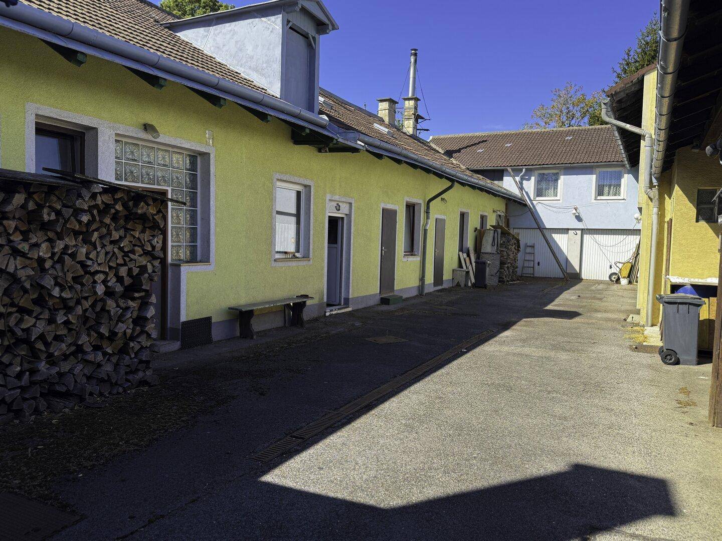 Sonniger Innenhof mit gelber Fassade, Holzstapel und Zugangstüren.