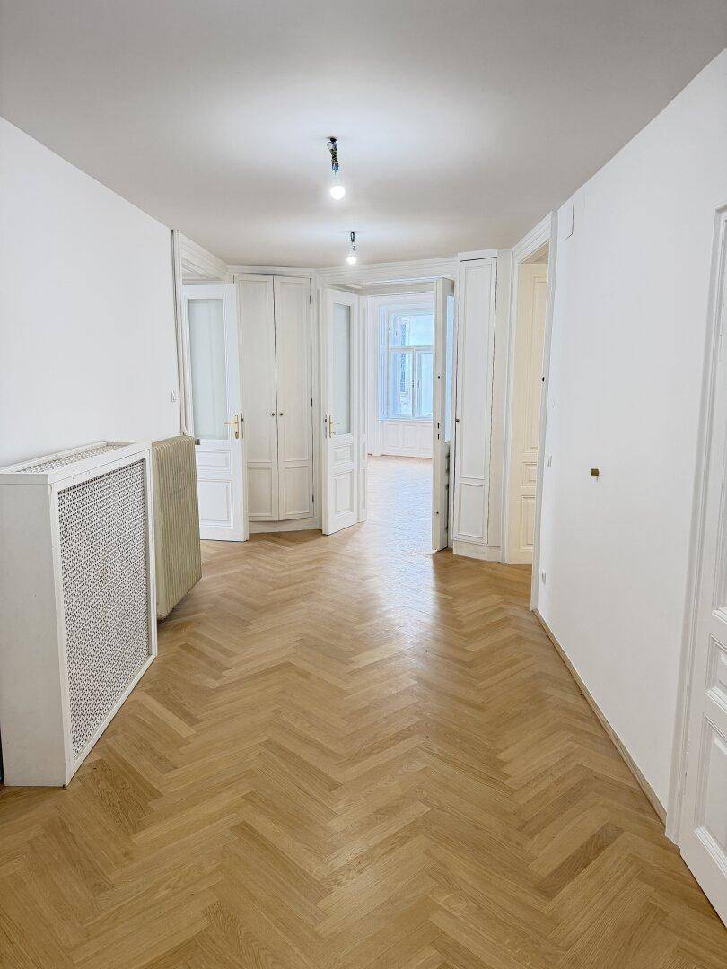 Langer Flur mit Fischgrätparkett und mehreren Türen, der zu weiteren Räumen und einem Erkerfenster führt.