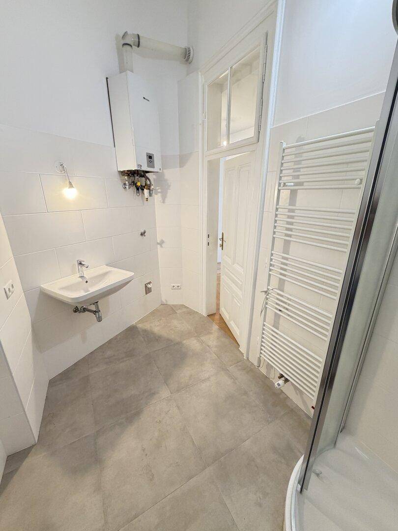 Helles Badezimmer mit weißen Fliesen, Duschkabine, Handtuchheizkörper und einem Wandwaschbecken.