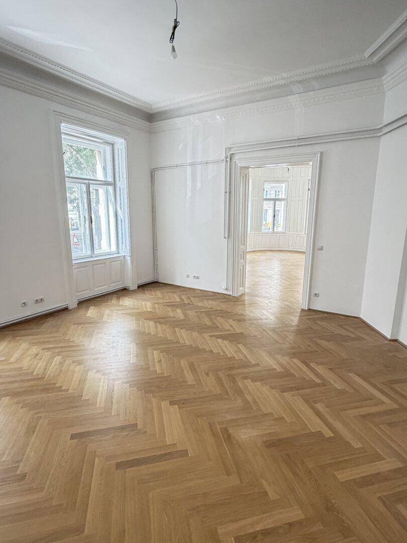 Helles Zimmer mit Fischgrätparkett, hohen Decken und einem großen Fenster, das den Blick nach draußen freigibt.