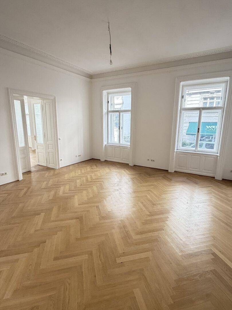 Geräumiges Zimmer mit Fischgrätparkett, hohen Decken und zwei großen Fenstern, die viel Helligkeit bieten.