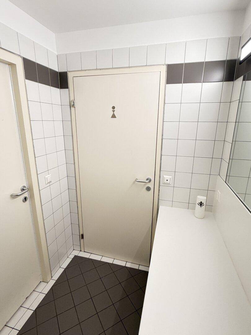 Toilettenbereich mit weißen Fliesen, dunklem Boden und einer Tür mit Piktogramm.