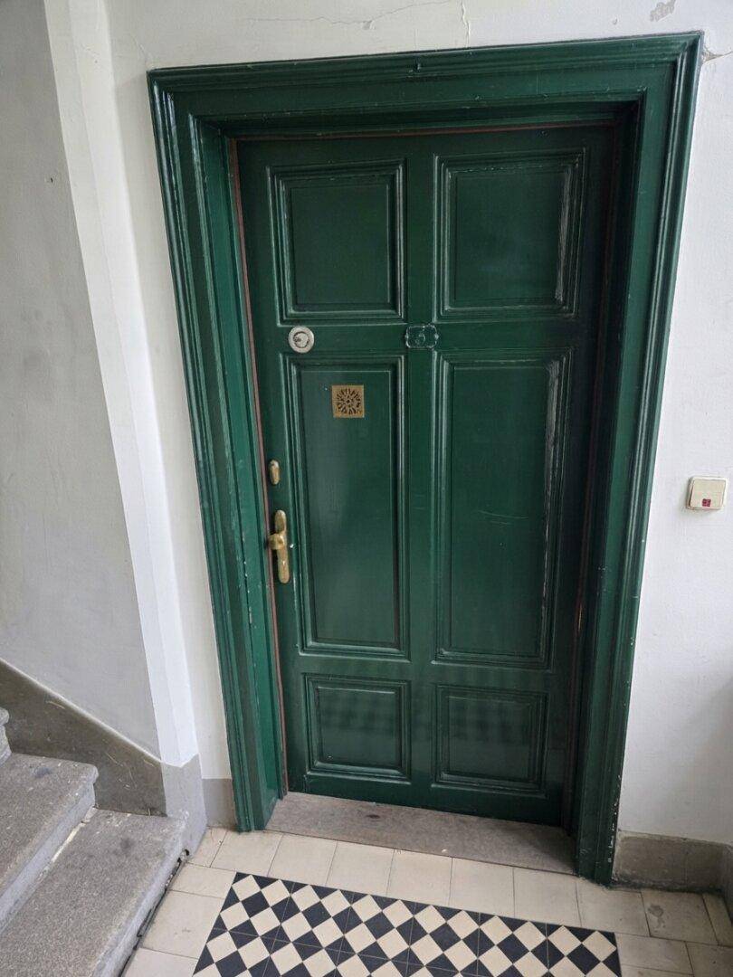 Dunkelgrüne Eingangstür mit traditionellem Kassettenmuster und schwarz-weißem Fliesenboden im Treppenhaus.