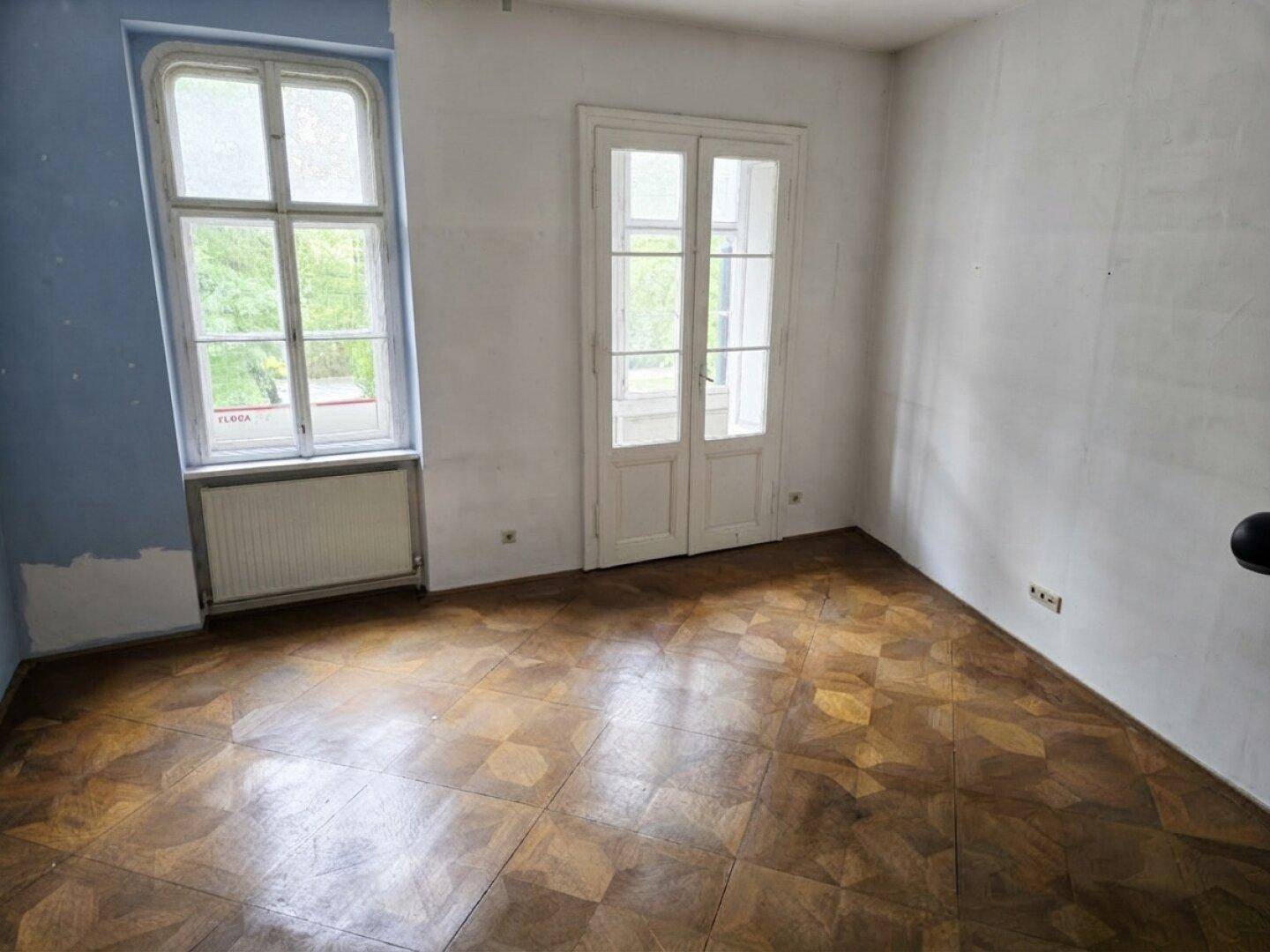 Lichtdurchfluteter Raum mit Parkettboden, großem Fenster und Flügeltüren, ideal für Wohnzwecke.