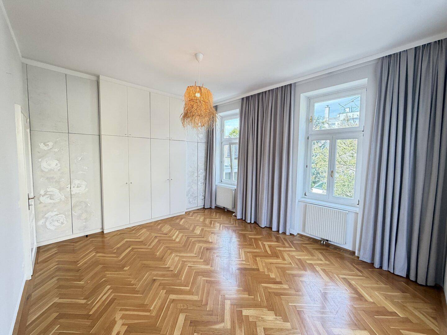Schlafzimmer mit Fischgrätparkett, Einbauschränken und großen Fenstern mit Vorhängen.