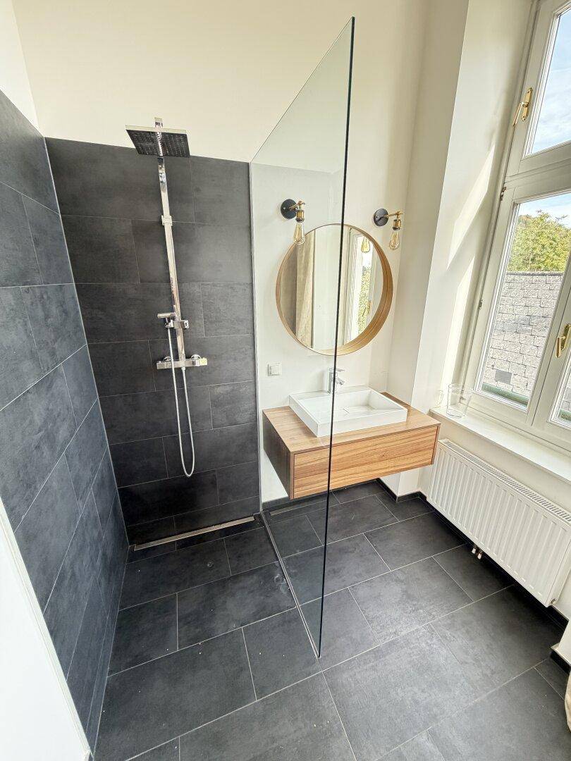 Modernes Badezimmer mit ebenerdiger Dusche, dunklen Fliesen und Holzwaschtisch.