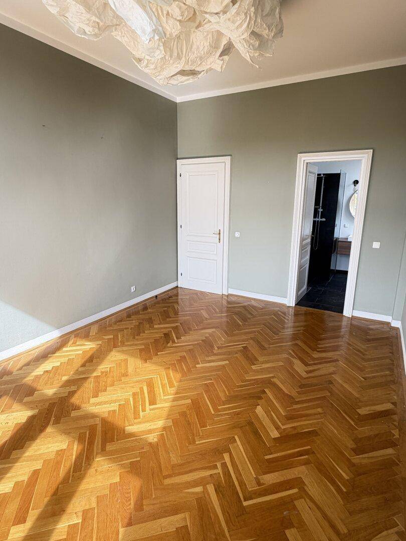 Geräumiges Zimmer mit Fischgrätparkett, hohen Decken und modernem Deckenlicht.