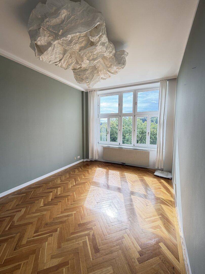 Helles Zimmer mit Fischgrätparkett, großen Fenstern und Blick ins Grüne.