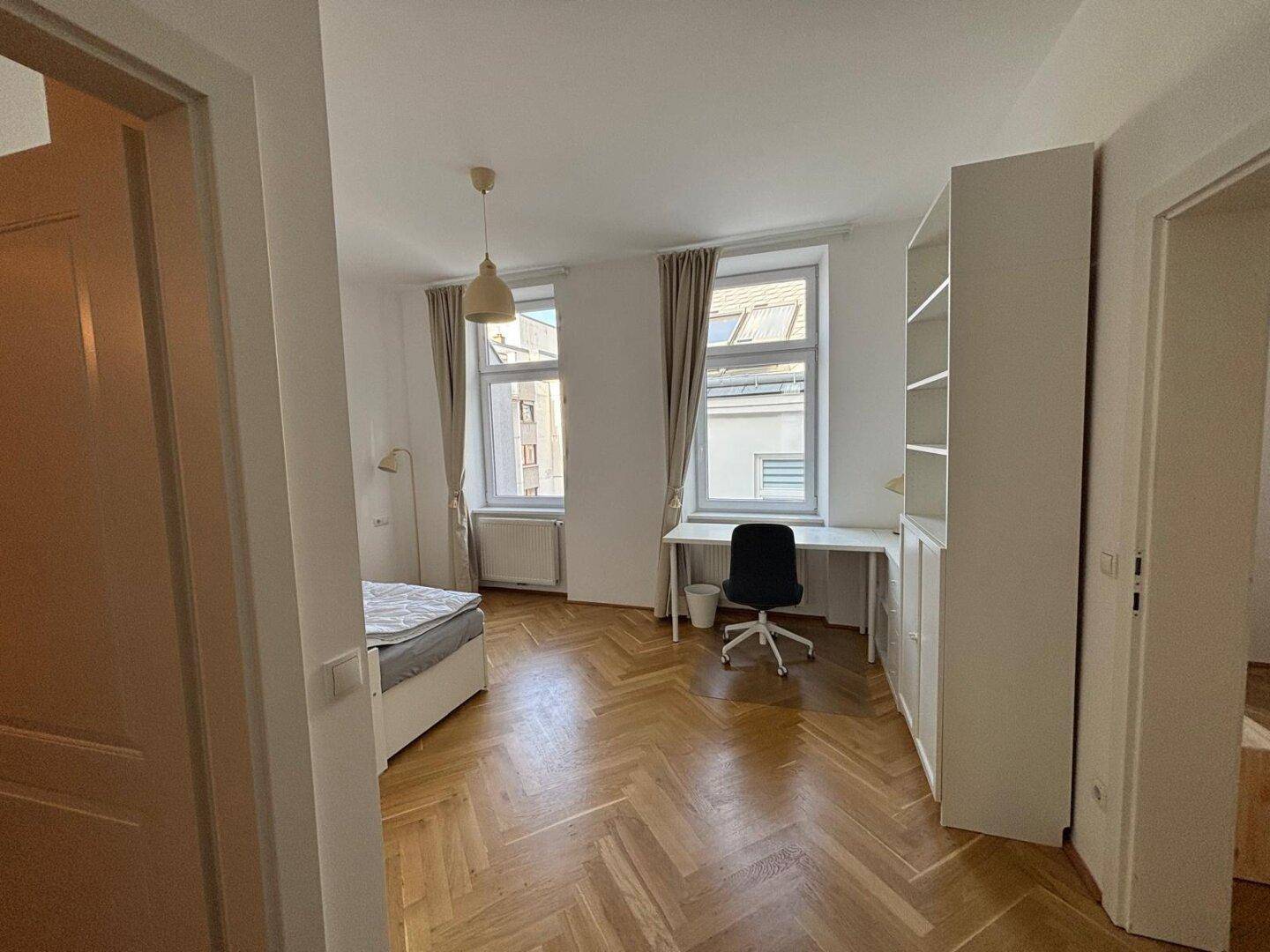 Schlafzimmer mit Parkettboden, Bett und Schreibtisch, gut beleuchtet durch große Fenster.