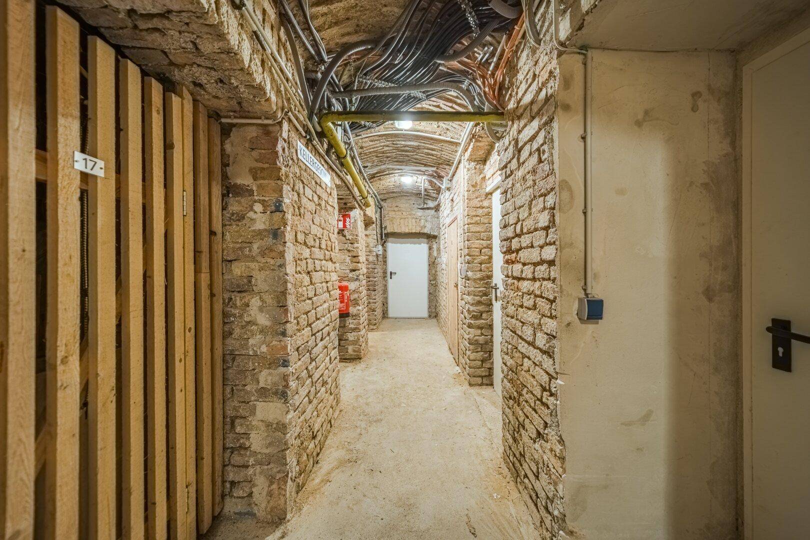 Historischer Kellergang mit Ziegelwänden, Gewölbedecke und Lagerabteilen, gut beleuchtet.