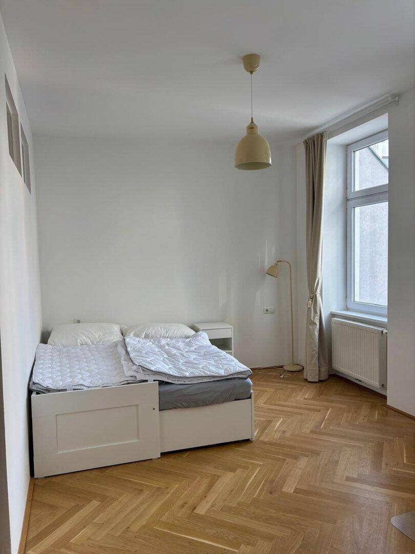 Gemütliches Schlafzimmer mit Parkettboden, Bett und heller Einrichtung, gut beleuchtet.