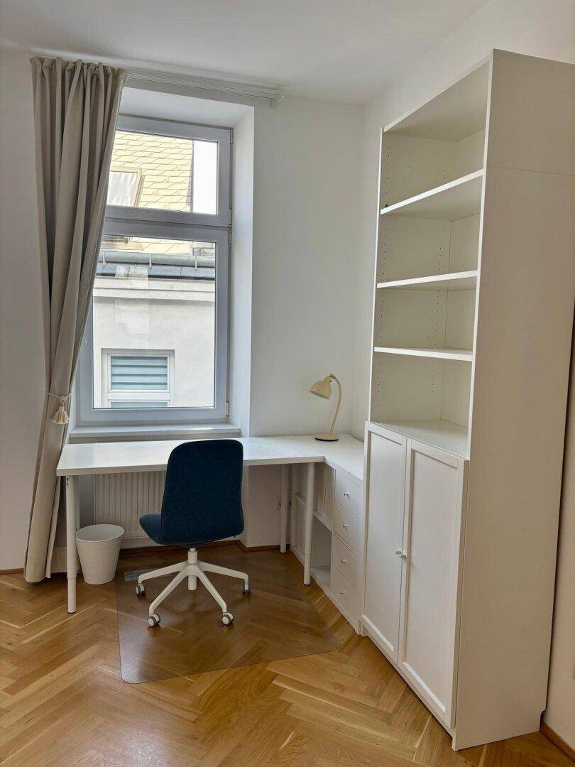 Arbeitsbereich mit Schreibtisch, Bürostuhl und Regal, gut beleuchtet durch ein Fenster.