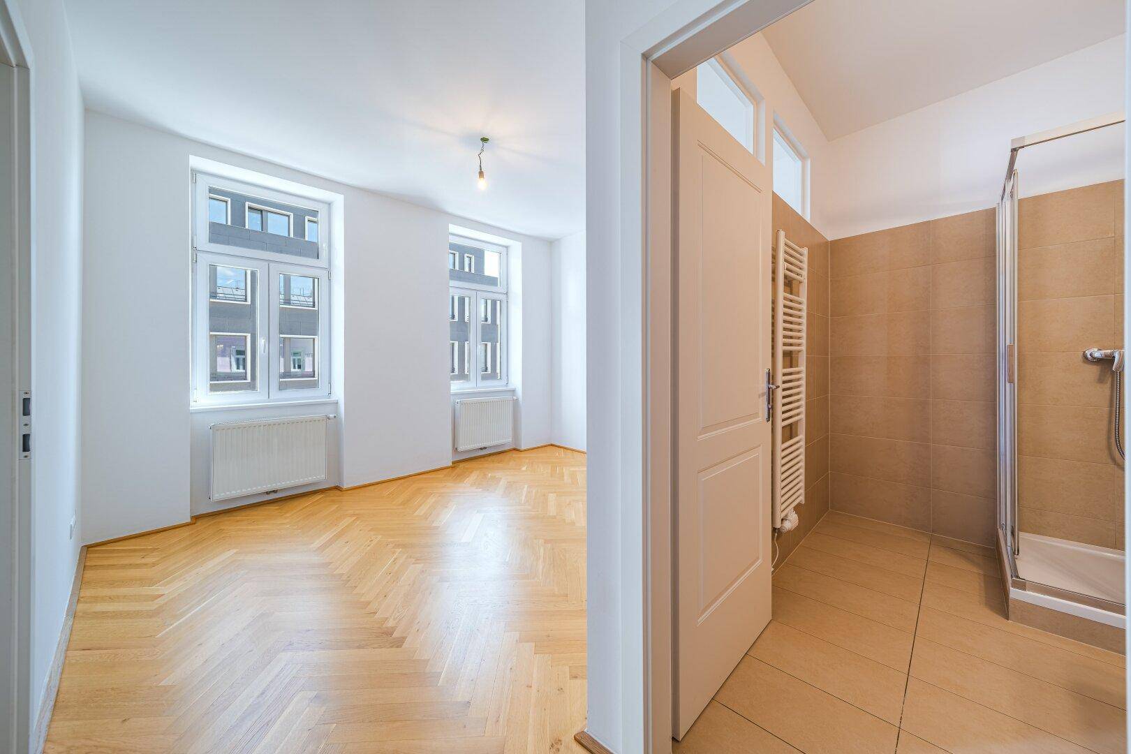 Heller Wohnraum mit Fischgrätparkett und Zugang zu einem Badezimmer mit Duschkabine.