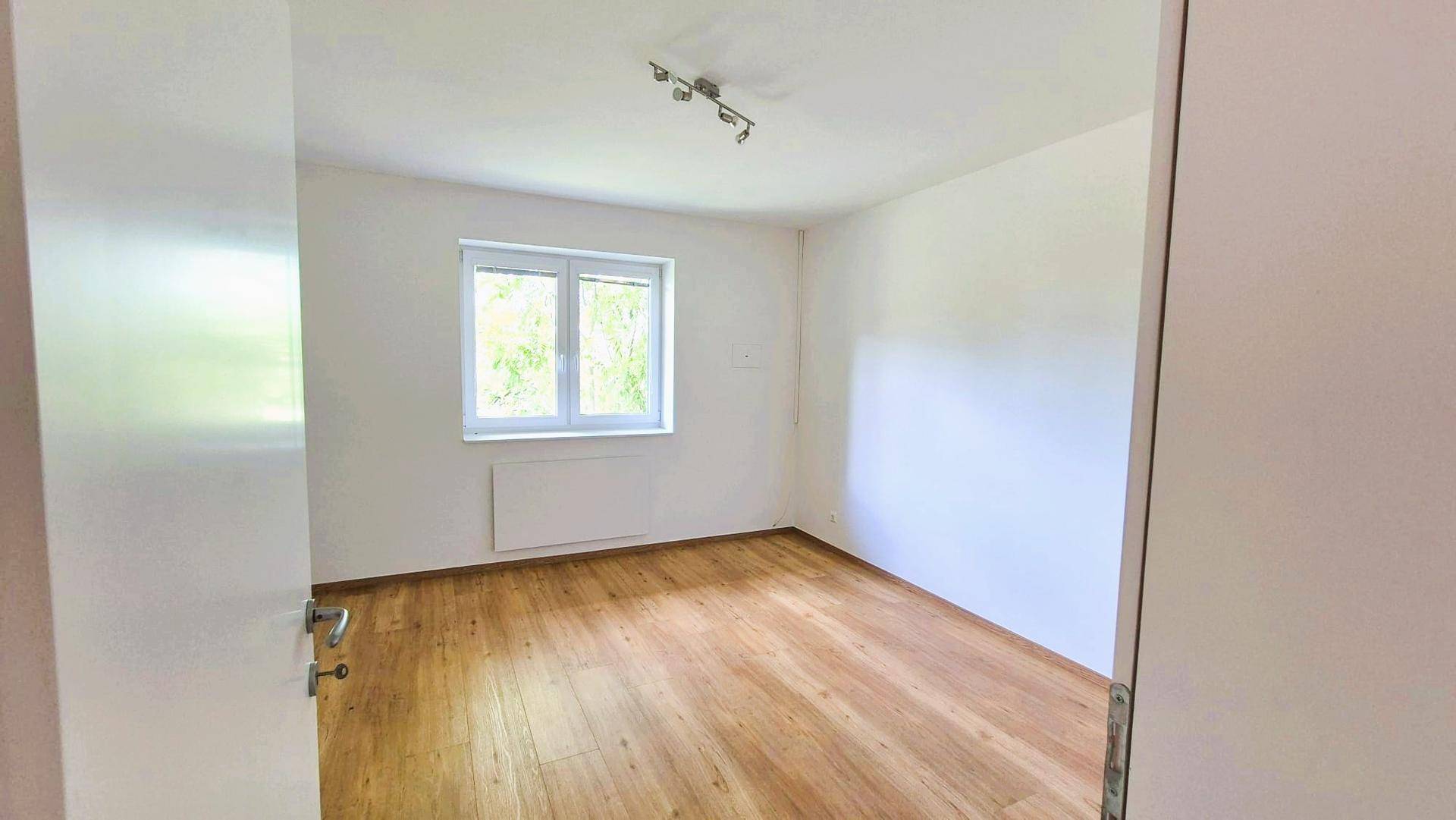 Helles, leeres Schlafzimmer mit Laminatboden und einem Fenster mit Blick ins Grüne.