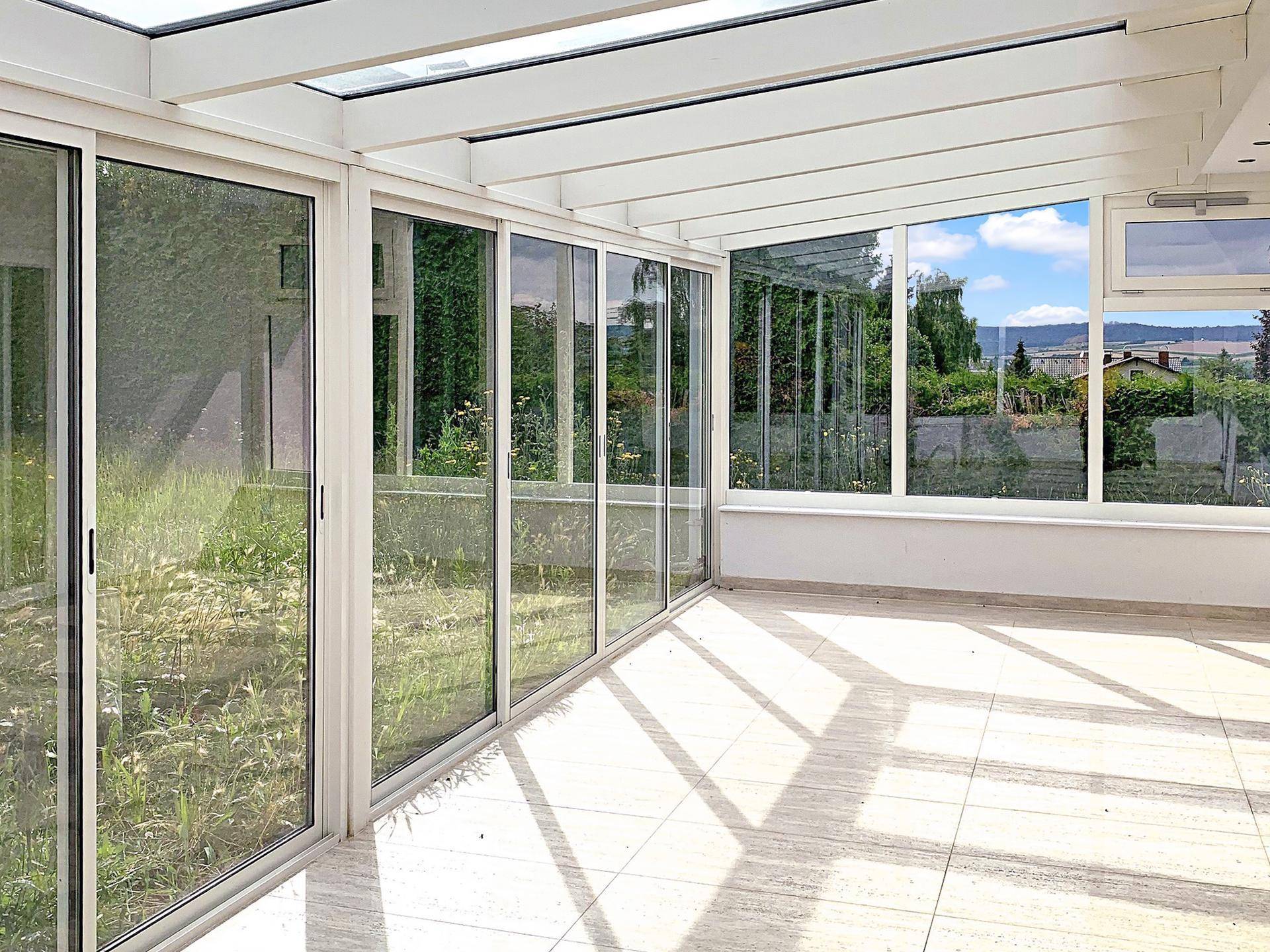Heller Wintergarten mit Fliesenboden, Glasdach und weitem Blick ins Grüne.