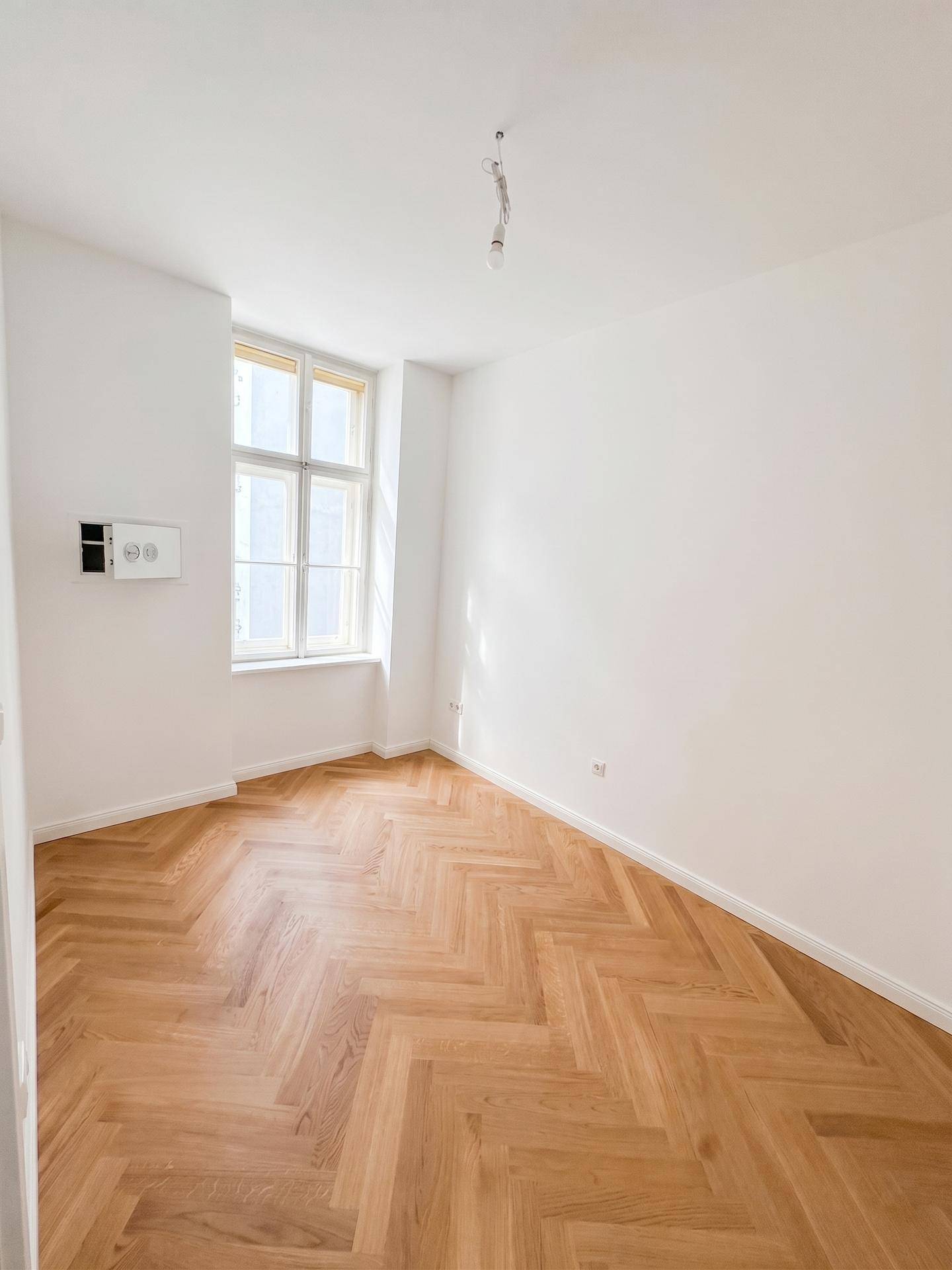 Geräumiges Zimmer mit Fischgrätparkett und großem Fenster, ideal als Schlaf- oder Arbeitszimmer.