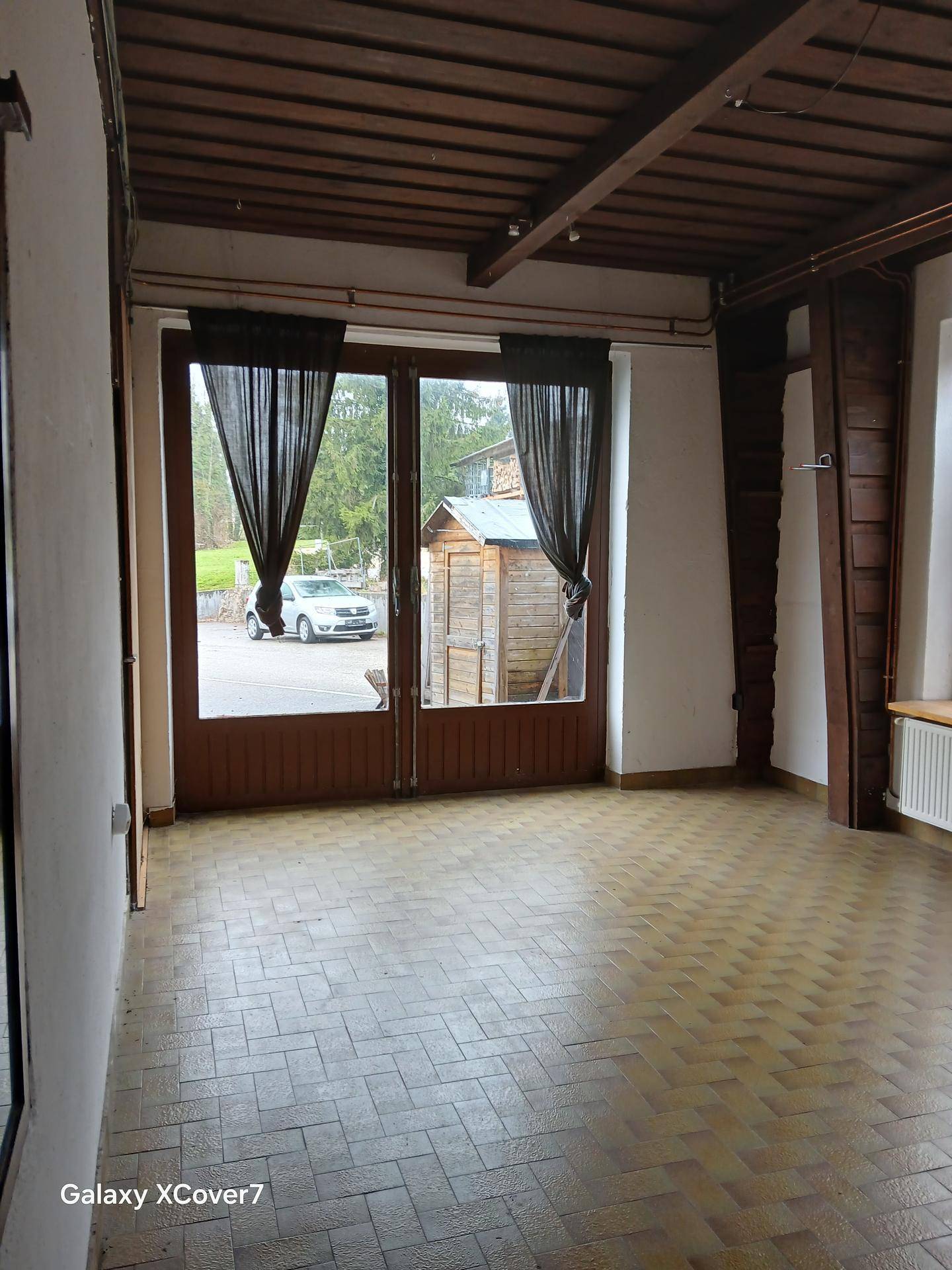 Innenraum mit Holzbalkendecke, Fliesenboden und großen Fenstertüren, die nach draußen zu einem Schuppen führen.