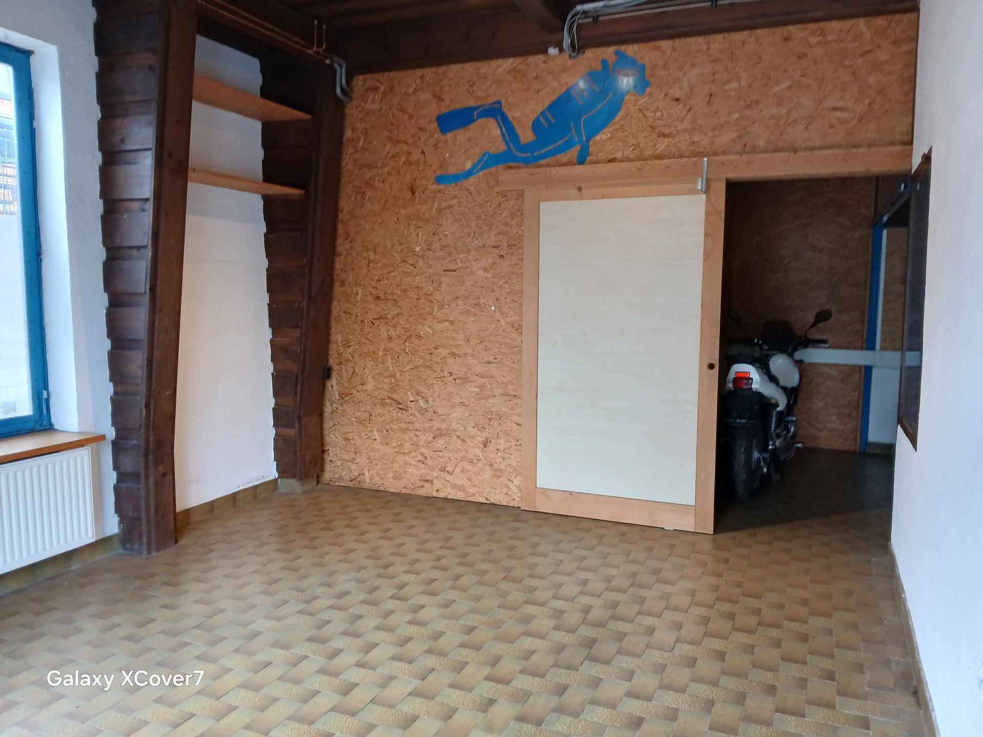 Innenraum mit OSB-Platten an der Wand, Holzbalken und einem Schiebetürzugang zu einem weiteren Bereich mit Motorrad.