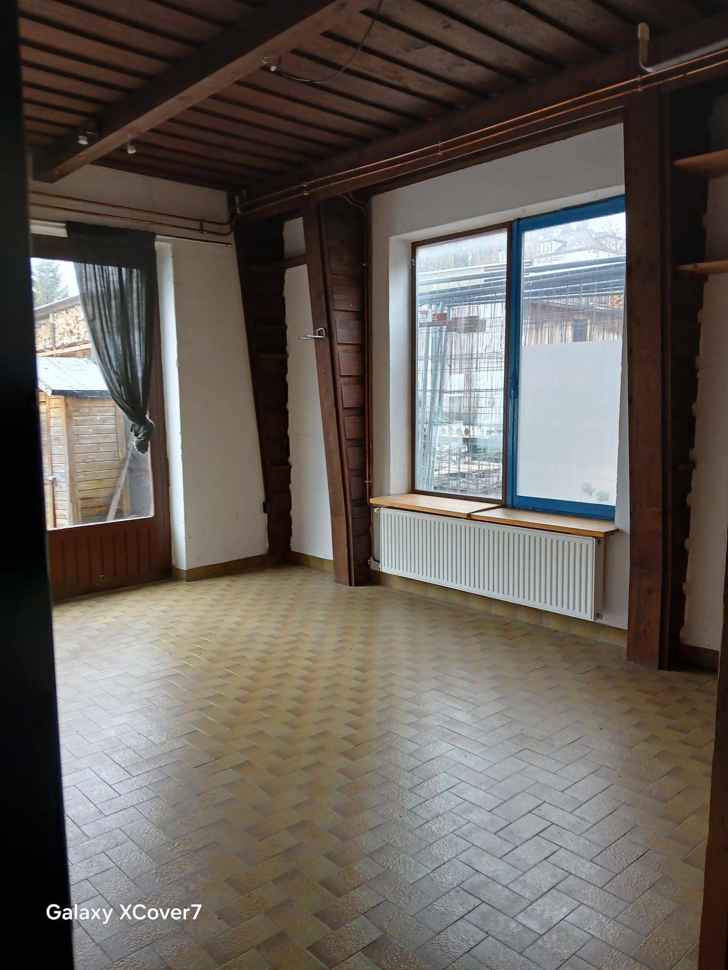 Geräumiger Innenraum mit sichtbaren Holzbalken, Fliesenboden und einem Heizkörper unter einem großen Fenster.