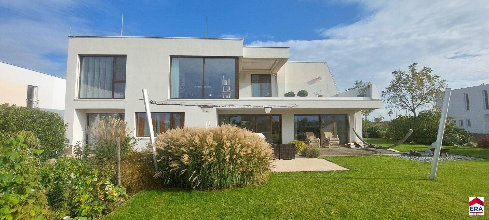 Moderne Villa mit großen Glasfronten, gepflegtem Garten und gemütlicher Terrasse unter blauem Himmel.