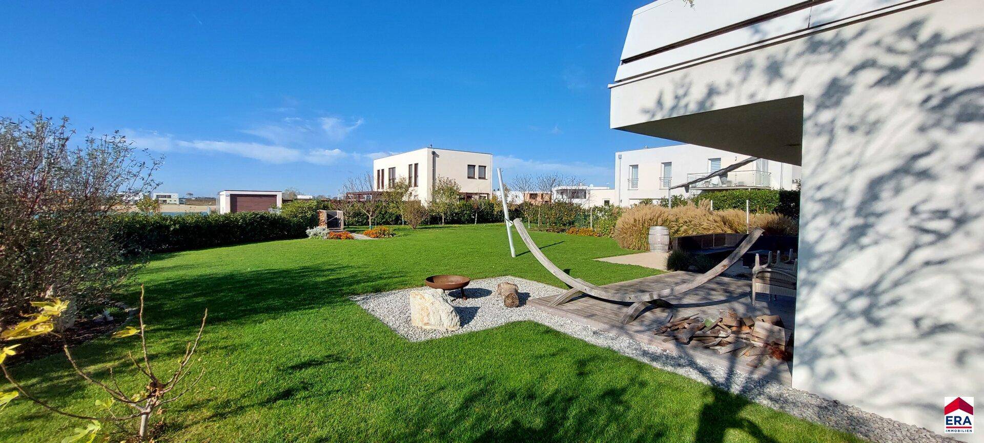 Weitläufiger Garten mit Rasenfläche, Feuerstelle und Liegemöglichkeit unter blauem Himmel.