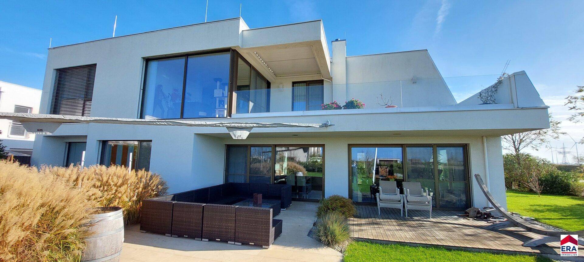 Moderne Villa mit großen Glasfronten, Terrasse und gepflegtem Garten unter blauem Himmel.