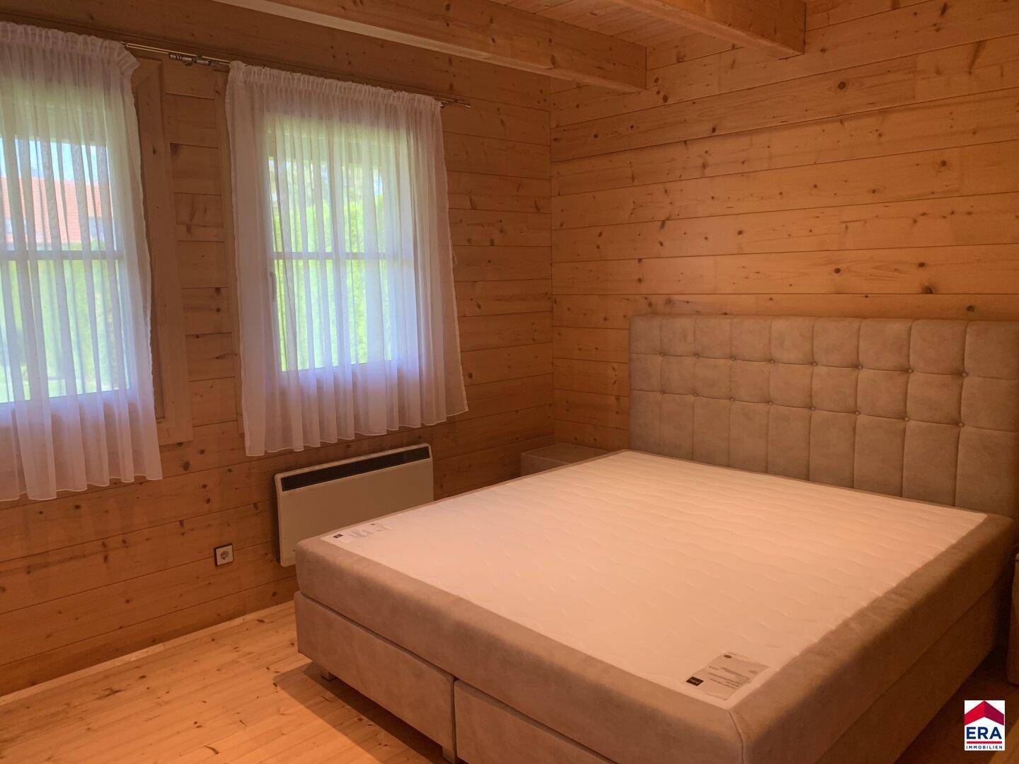Helles Schlafzimmer mit Holzwänden, Doppelbett und zwei Fenstern.