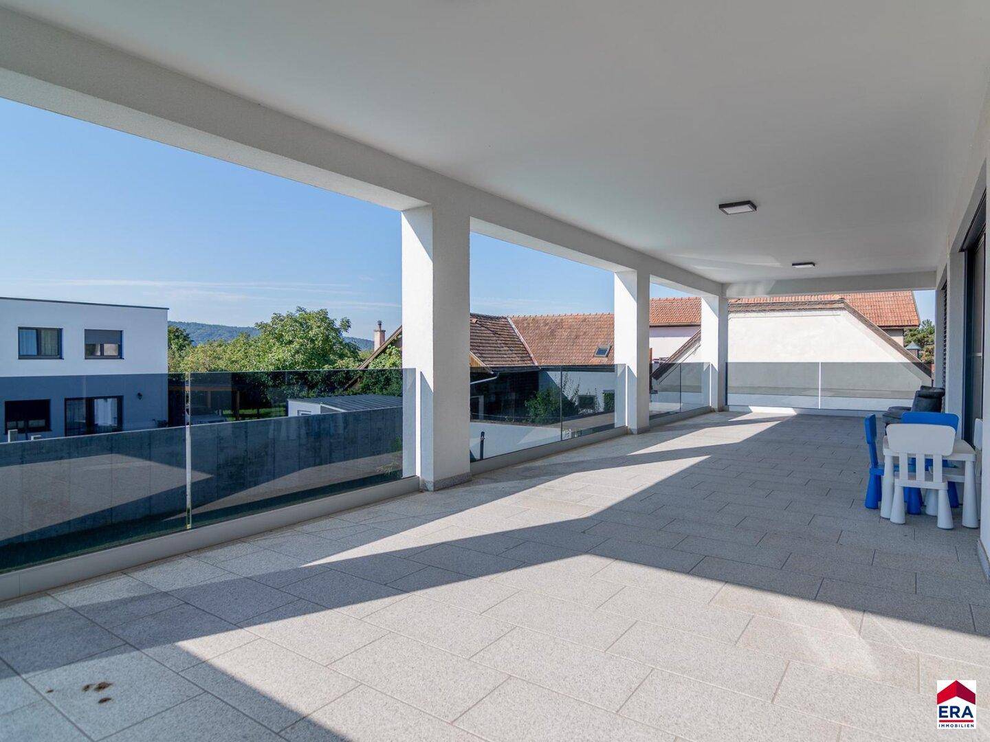 Weitläufige überdachte Terrasse mit Glasgeländer und Blick auf die grüne Umgebung.