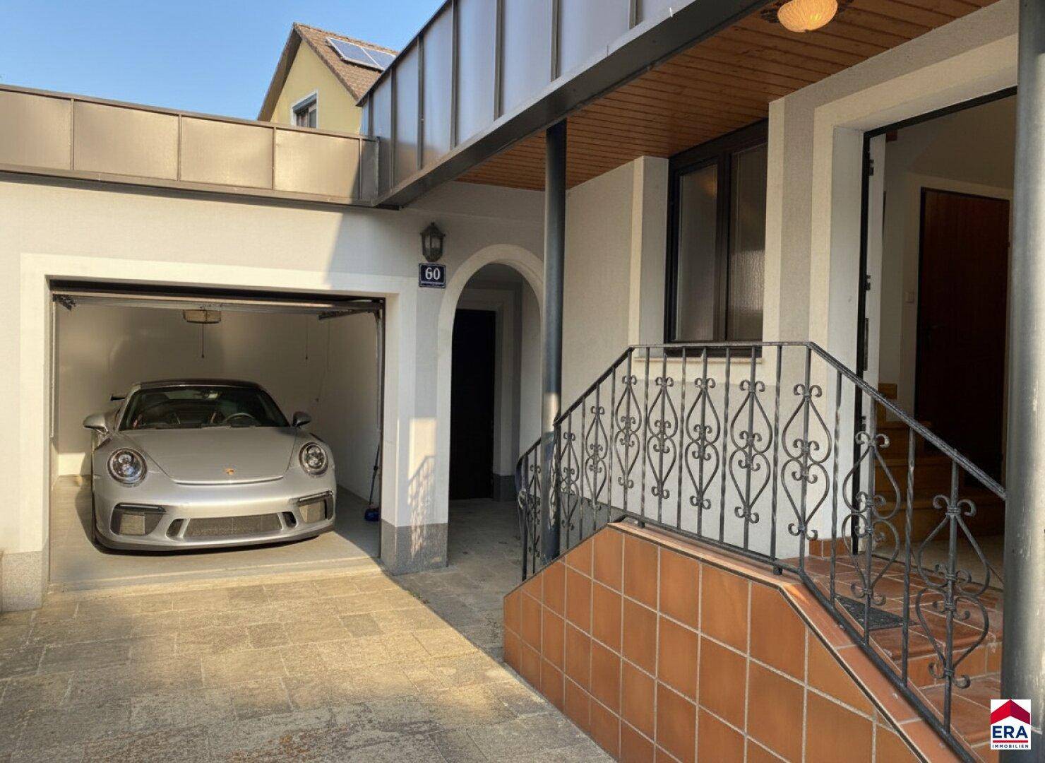 Eingangsbereich des Hauses mit Treppe, schmiedeeisernem Geländer und einer Garage mit geparktem Sportwagen.