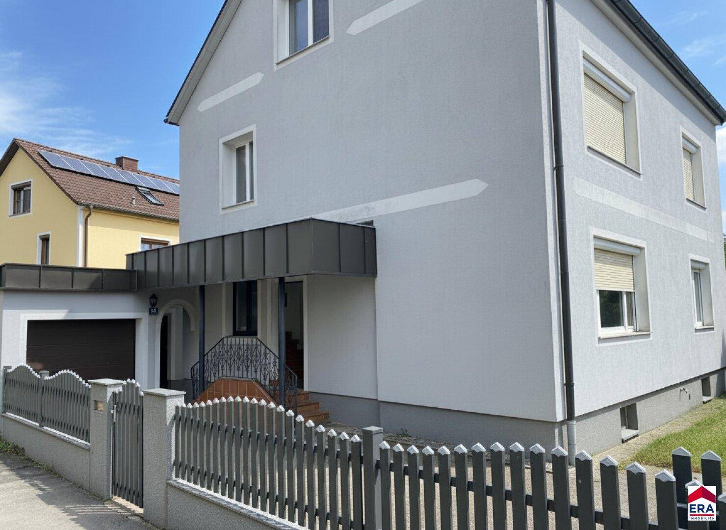 Vorderansicht des Hauses mit grauer Fassade, Rollläden und einem gepflegten Vorgarten mit Zaun.
