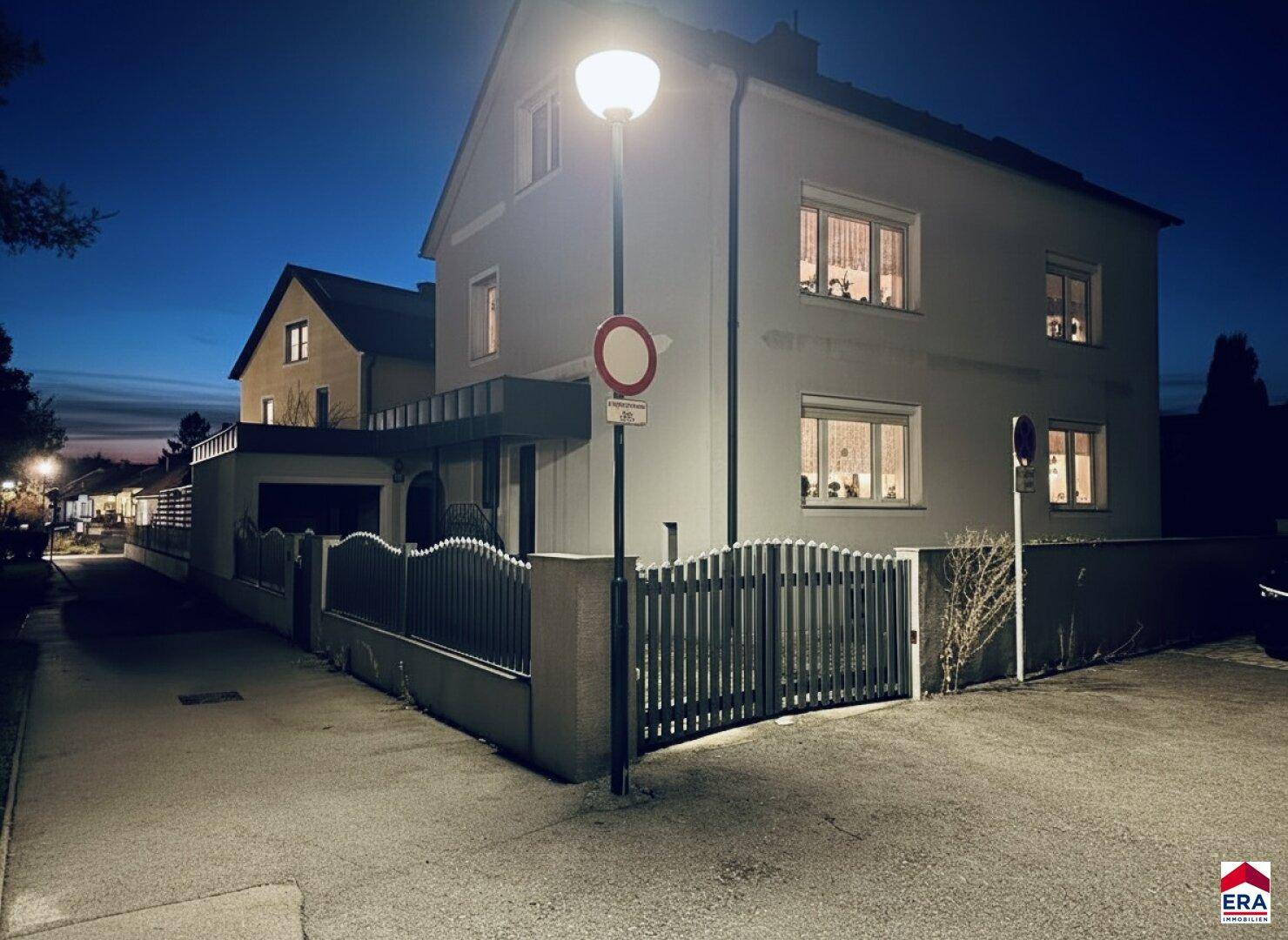 Einfamilienhaus bei Nacht mit beleuchteten Fenstern und einem Laternenpfahl vor dem Grundstück.