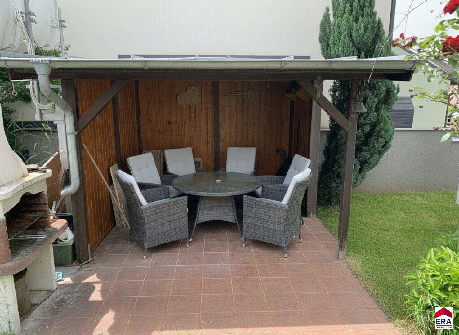 Überdachter Sitzbereich im Garten mit Rattanmöbeln, Esstisch und gemauertem Grill.