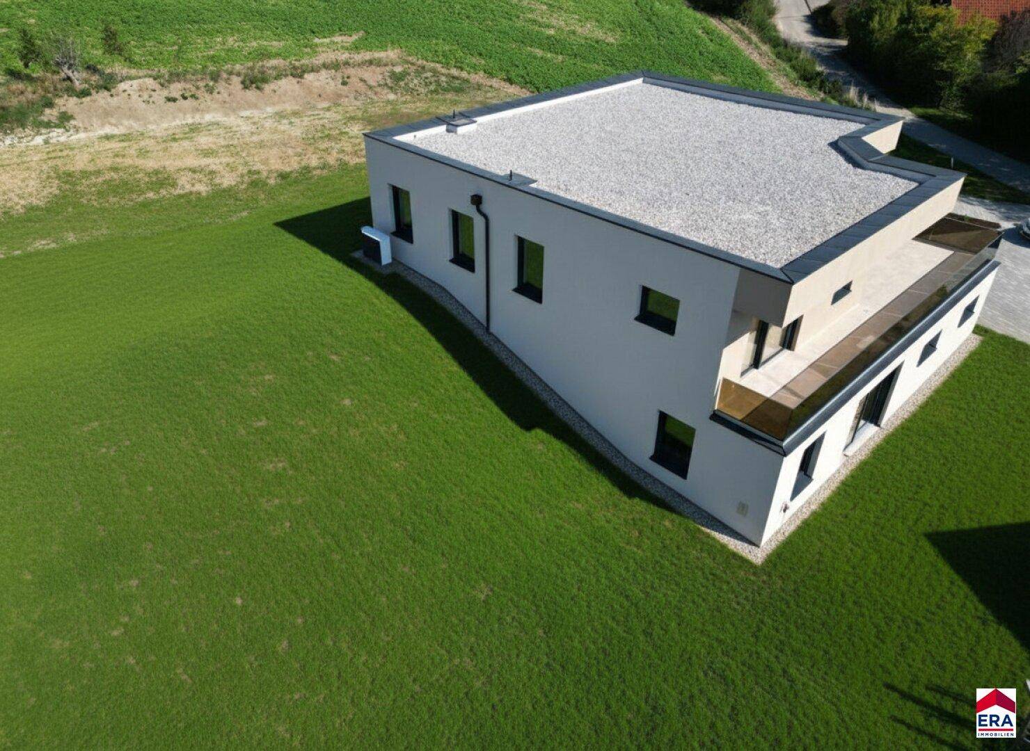 Weitläufige Luftaufnahme des modernen Neubaus mit Flachdach, umgeben von grünen Hügeln und Feldern.
