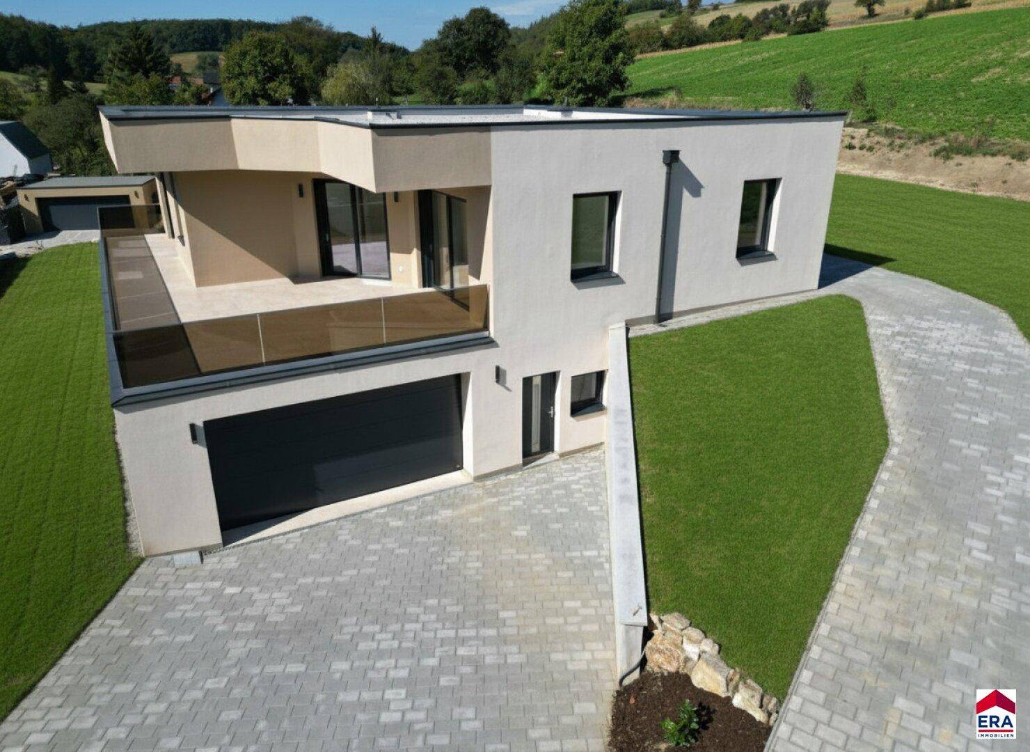 Moderne Hausfassade mit gepflasterter Auffahrt, Garage und weitläufigen Rasenflächen in ländlicher Umgebung.