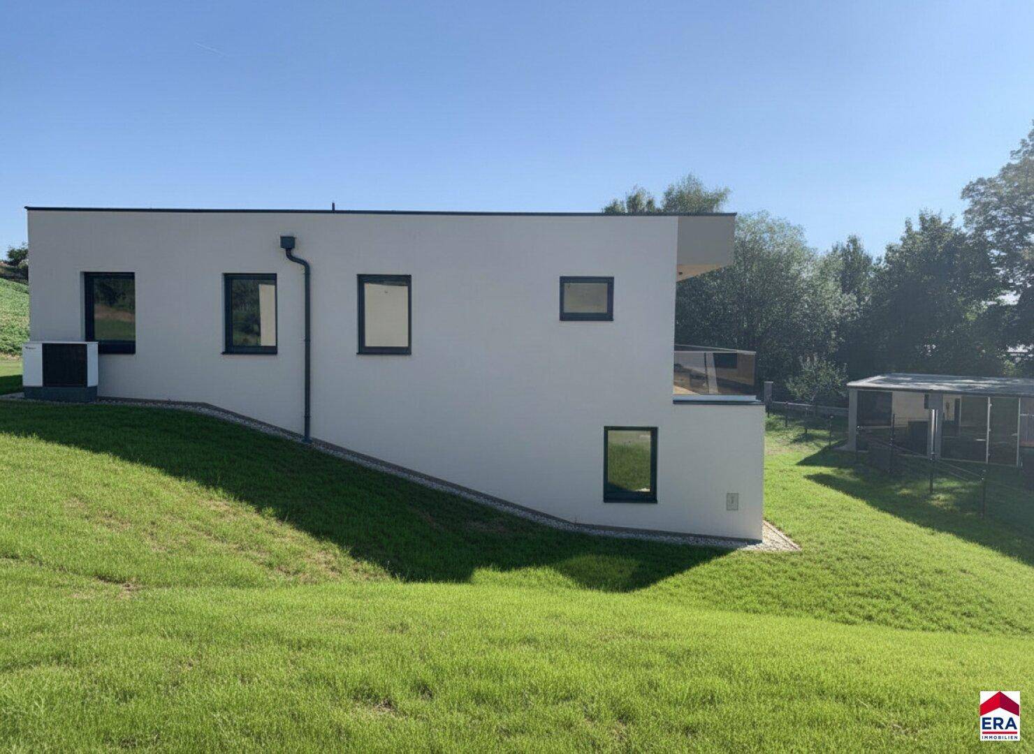 Seitenansicht des modernen Hauses mit weißer Fassade und grüner Hanglage unter blauem Himmel.