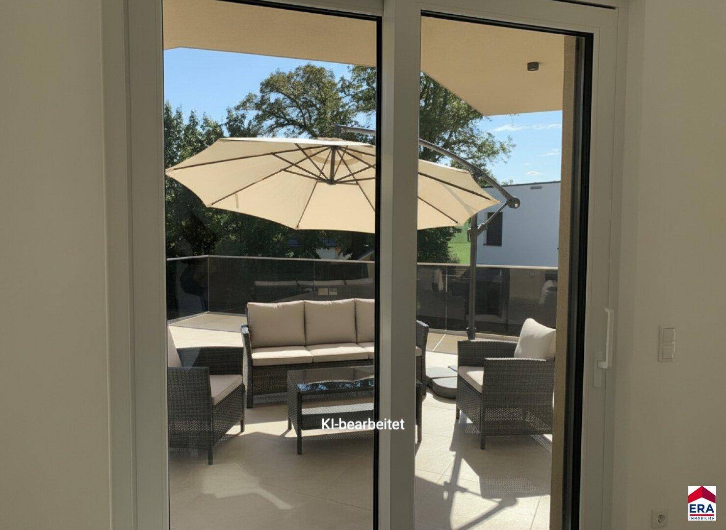 Blick vom hellen Innenraum durch große Schiebetüren auf die möblierte Terrasse mit Sonnenschirm.