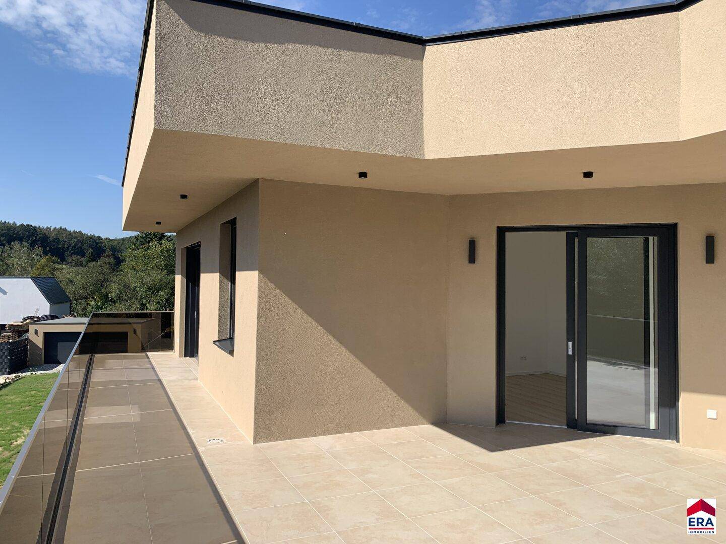 Geräumige, unmöblierte Terrasse mit Fliesenboden und Glasgeländer, bietet weiten Blick ins Grüne.