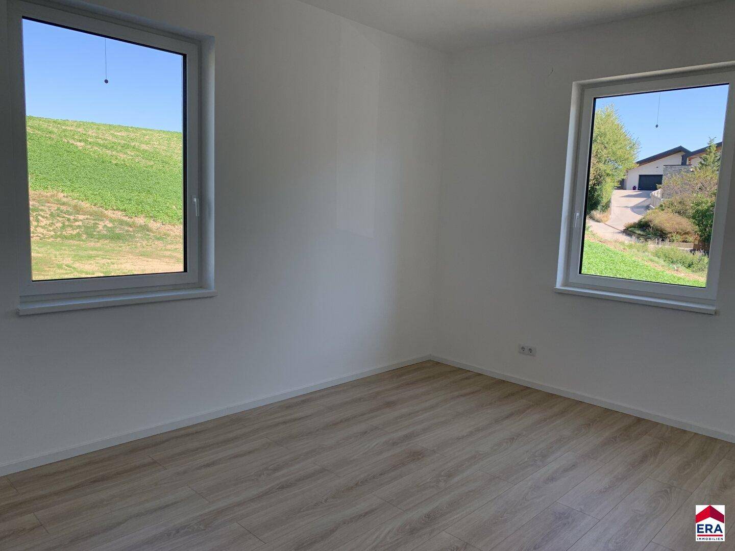 Helles, leeres Schlafzimmer mit Laminatboden und zwei Fenstern, die einen grünen Ausblick bieten.
